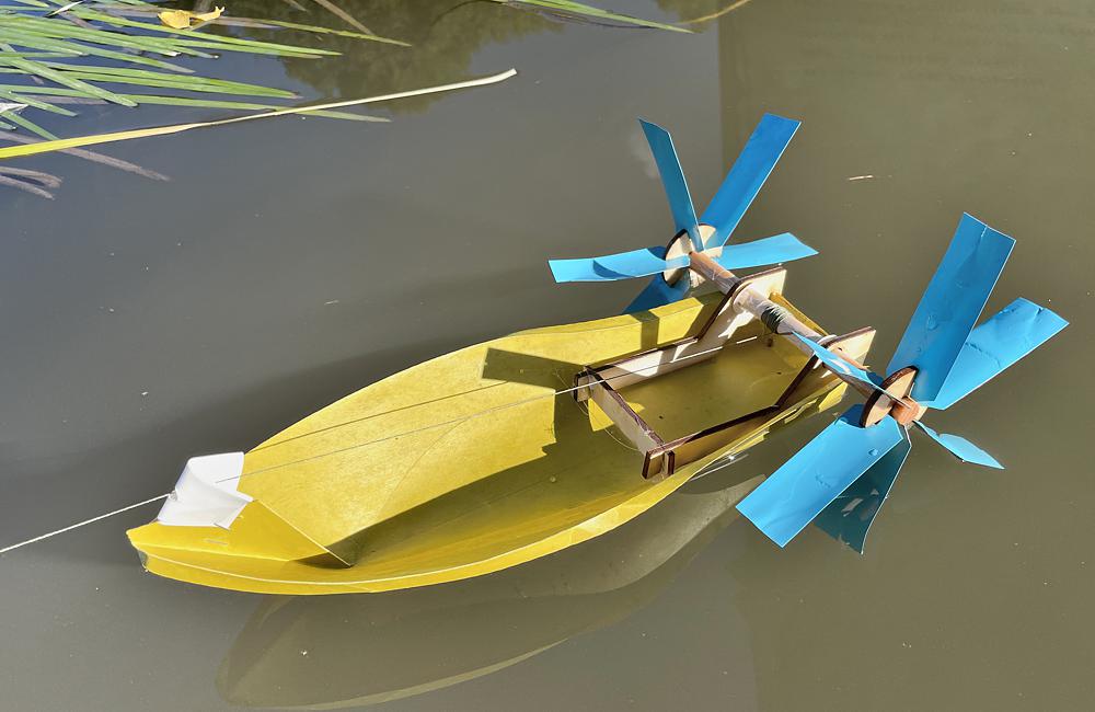 Detailansicht eines Schwimmkörpers zur Untersuchung der Wasserqualität, er hat die Form eines kleinen Bootes. Der Rumpf ist gelb, hinten sind zwei blaue Schaufelräder angebracht, die mit einer Schnur gesteuert werden können.
