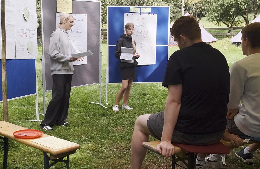 Auf einer Wiese stehen zwei jugendliche Moderatorinnen und halten ihre Vortragspapiere in der Hand. Hinter ihnen sind blaue und graue Stellwände mit großen Papierbögen. Vor ihnen sitzen die Zuhörer auf Holzbänken.