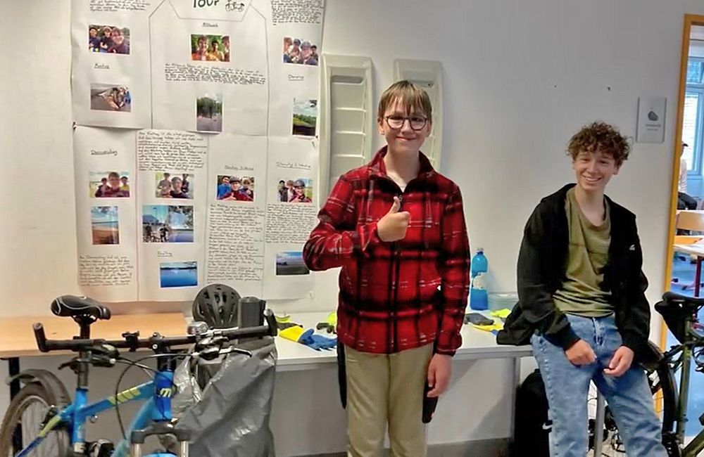 Präsentation des Projektes „Fahrradtour“. 2 Teilnehmer haben ihre Fahrräder und einen Teil ihrer Ausrüstung mitgebracht. An der Wand hinter ihnen haben sie auf großen Papierbögen mit Fotos und Fakten zu ihrer Fahrradtour aufgehängt.
