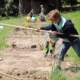 Sechs Schulkinder bereiten ein Beet im Schulgarten vor. Im Vordergrund lockert ein Junge die Erde mit einer langen Hacke auf, im Hintergrund knien die anderen Kinder.