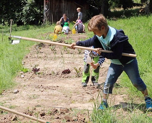 Sechs Schulkinder bereiten ein Beet im Schulgarten vor. Im Vordergrund lockert ein Junge die Erde mit einer langen Hacke auf, im Hintergrund knien die anderen Kinder.