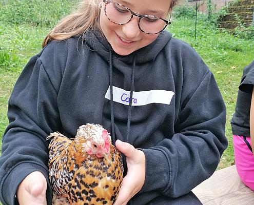 Ein Mädchen mit blauem Kapuzenpulli hält in seinen Händen ein junges Huhn. Das Huhn hat hellbraune Federn mit schwarzen Spitzen. Das Mädchen hat ein Namenschild „Clara“ auf der Brust befestigt.