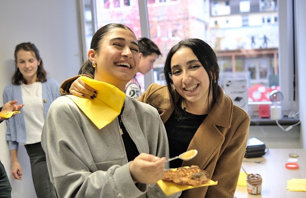 Die Jugendlichen haben Waffeln gebacken. 2 fröhliche dunkelhaarige Frauen probieren eine Waffel mit heller Creme auf gelben Servietten.