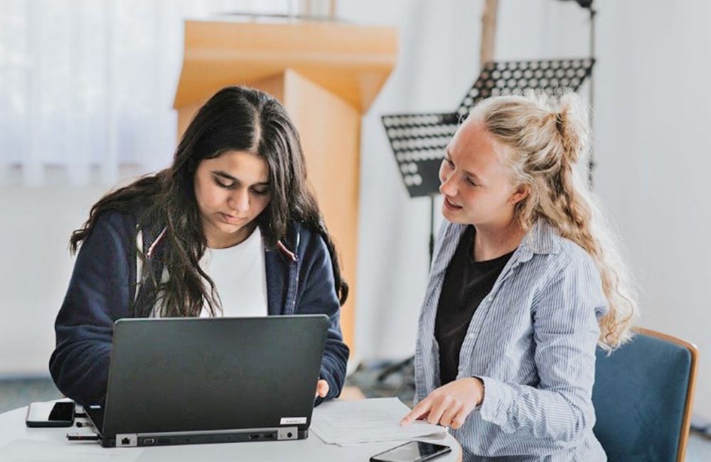 2 junge Frauen sitzen an einem Tisch. Die dunkelhaarige arbeitet konzentriert an einem Laptop, die hellhaarige erklärt ihr etwas.