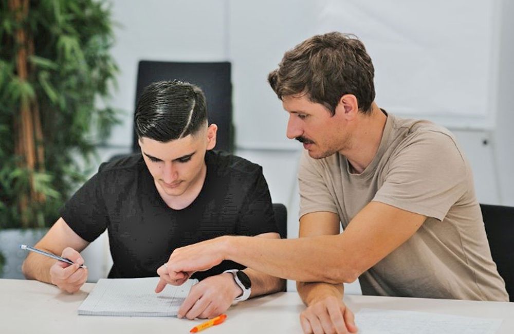 2 junge Männer sitzen an einem Tisch und bearbeiten gemeinsam einen Text auf Papier.