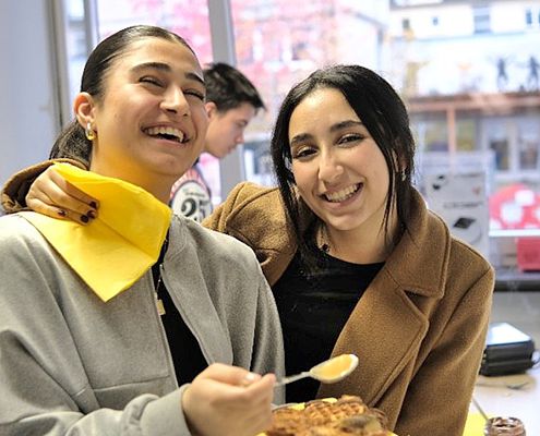 Die Jugendlichen haben Waffeln gebacken. 2 fröhliche dunkelhaarige Frauen probieren eine Waffel mit heller Creme auf gelben Servietten.