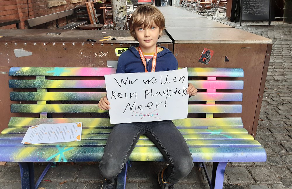 Ein Junge sitzt auf einer Bank, die mit bunter Farbe besprüht ist. Er blickt in die Kamera und hält ein Schild, auf dem steht: „Wir wollen kein Plastick Meer!“