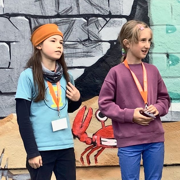 Zwei Mädchen stehen nebeneinander und blicken gespannt nach rechts. Sie haben Namenschilder an orangefarbenen Bändern umgehängt und blicken zur Interviewerin. Die Wand im Hintergrund ist bunt bemalt.